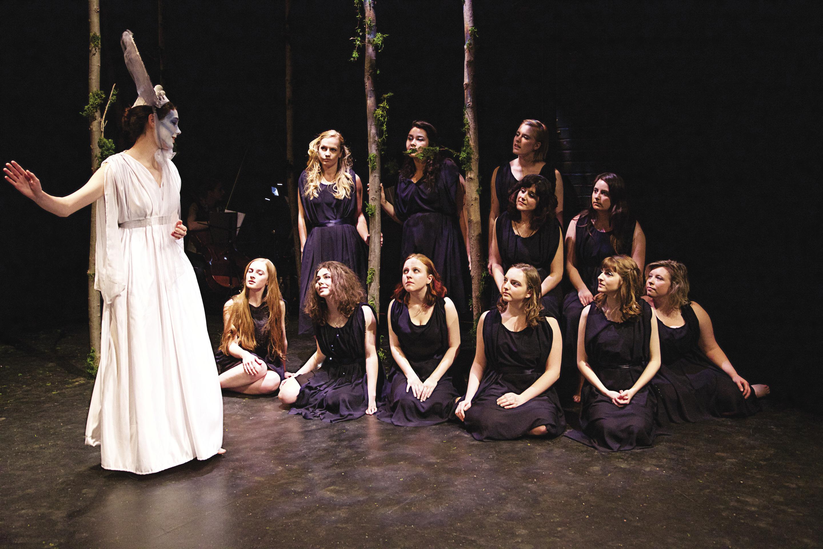 A woman in white walks past a group of women in black, kneeling on a stage